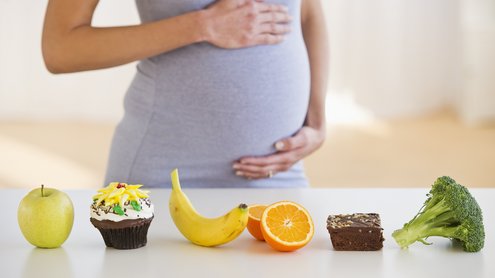 Schwangere steht hinter einem Tisch und h&auml;lt ihren Bauch. Auf dem Tisch steht ein Apfel, ein Mussif, eine Banane, eine Orange, ein Brownie und ein Brokkoli.